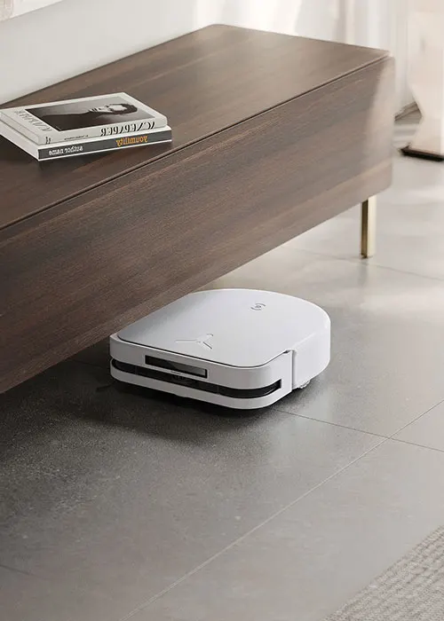 Un tavolino di legno marrone con libri sopra e un robot aspirapolvere bianco sul pavimento grigio sottostante.