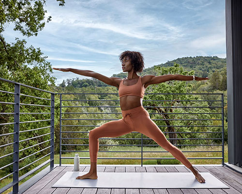 Donna in tenuta sportiva arancione fa yoga su un tappetino bianco su un balcone con ringhiera metallica, sullo sfondo alberi e cielo.