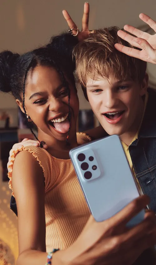 Due giovani sorridono e posano per una foto con un telefono cellulare grigio chiaro. La ragazza tiene il telefono mentre il ragazzo saluta.