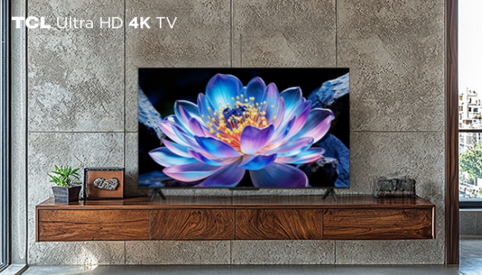 Un televisore TCL Ultra HD 4K TV posizionato su un mobile sospeso in legno venato, davanti a una parete in cemento grezzo. Sullo schermo è visualizzato il primo piano di un fiore di loto luminoso dalle sfumature blu e viola.