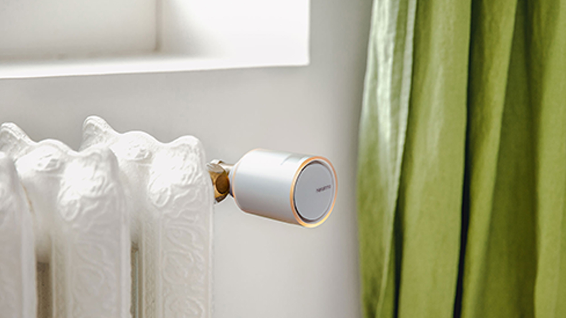 Termostato smart su termosifone bianco, con finestra e tenda verde sullo sfondo.