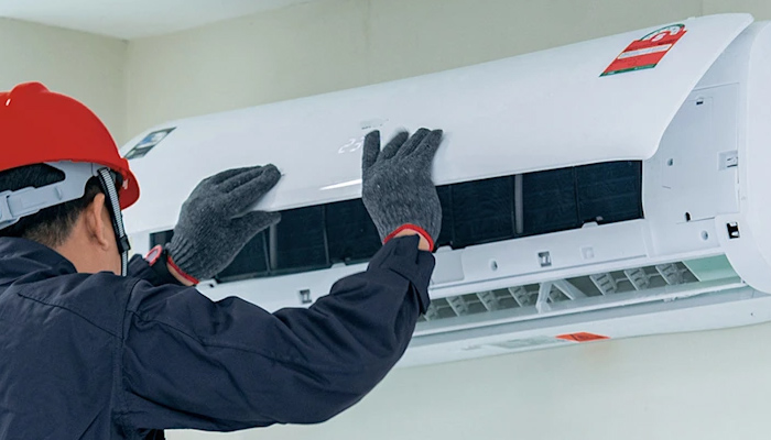Operaio con casco rosso che ripara un condizionatore bianco fissato al muro.