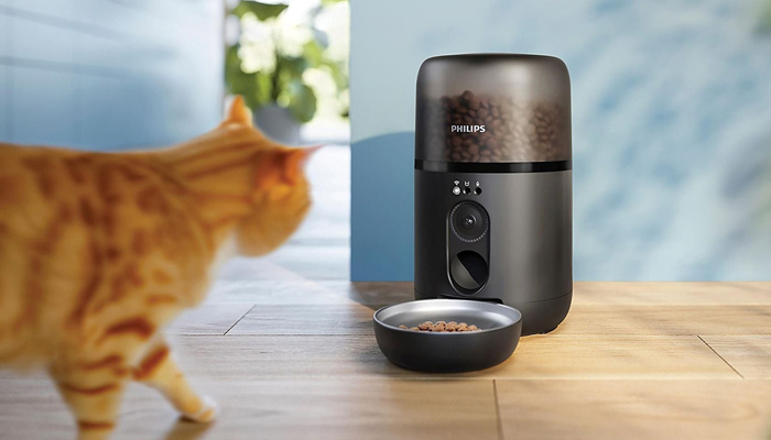 Un gatto soriano arancione si avvicina a un distributore automatico di cibo smart della Philips. Il dispositivo è nero con un contenitore trasparente pieno di croccantini e una ciotola coordinata posizionata su un pavimento in legno chiaro.
