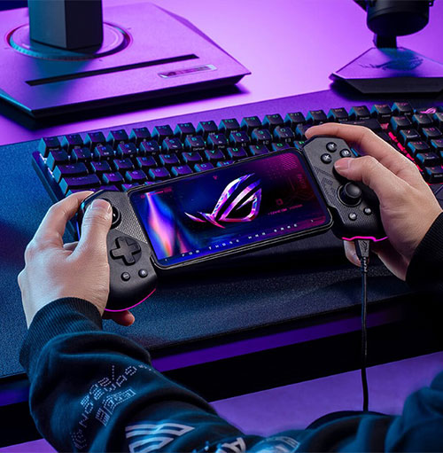 Mani che tengono un telefono con controller di gioco in una stanza illuminata di viola. Tastiera e monitor sullo sfondo.