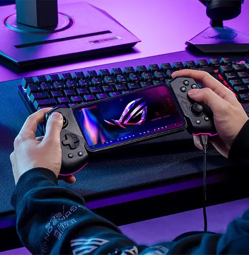 Mani che tengono un telefono con controller di gioco in una stanza illuminata di viola. Tastiera e monitor sullo sfondo.