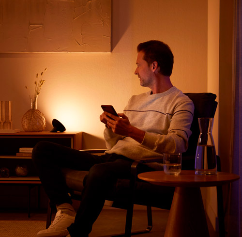 Uomo seduto su una sedia, tiene un cellulare. Accanto, tavolino con brocca d'acqua. In stanza, mobile con vaso e speaker.