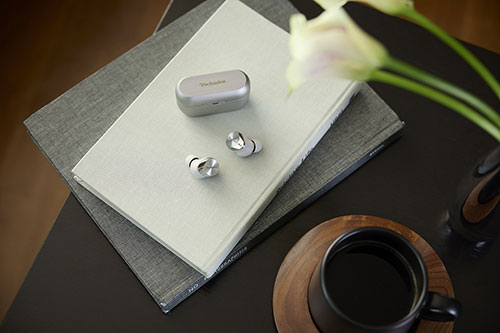 Auricolari e custodia grigi su libri impilati. Tazza nera piena su piattino di legno. Fiori bianchi in vaso.