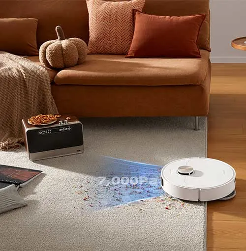 Divano marrone con cuscini, robot aspirapolvere bianco su tappeto chiaro. Radio con snack. Ambiente domestico.