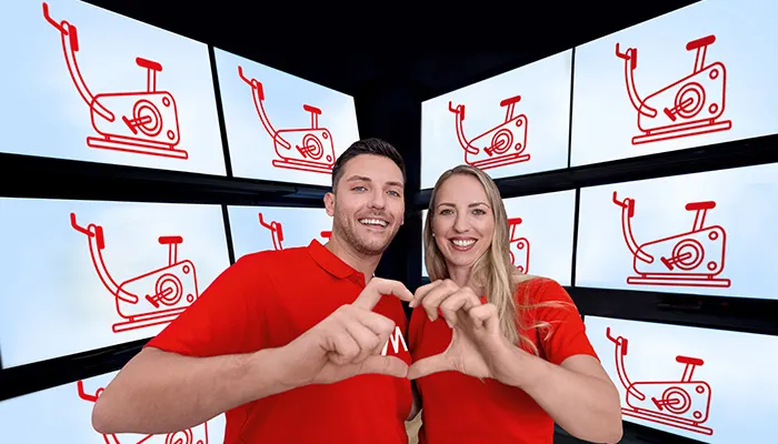 Uomo e donna sorridono, mani a forma di cuore, magliette rosse. Sfondo: schermi con icone rosse di cyclette.