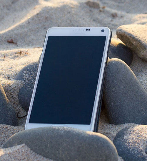 Telefono bianco con schermo scuro, appoggiato su sabbia e pietre grigie.