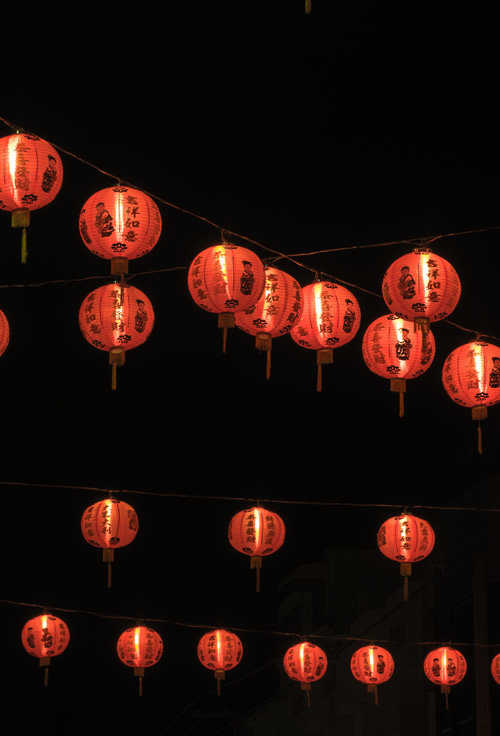 File di lanterne rosse illuminate con scritte nere sospese su fili, contro uno sfondo scuro notturno.