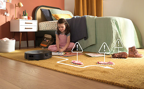 Ragazza seduta sul tappeto con robot aspirapolvere. Letti, scarpe e segnali di avvertimento visibili.