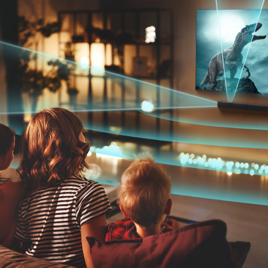 Tre bambini guardano un dinosauro su uno schermo TV. L'immagine è proiettata con raggi blu in una stanza buia.