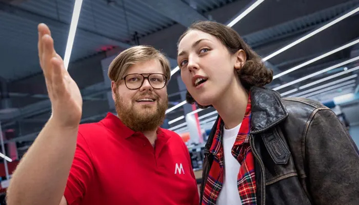 Uomo in polo rossa con logo saluta, donna con giacca in pelle guarda in alto. Luci al neon sullo sfondo.
