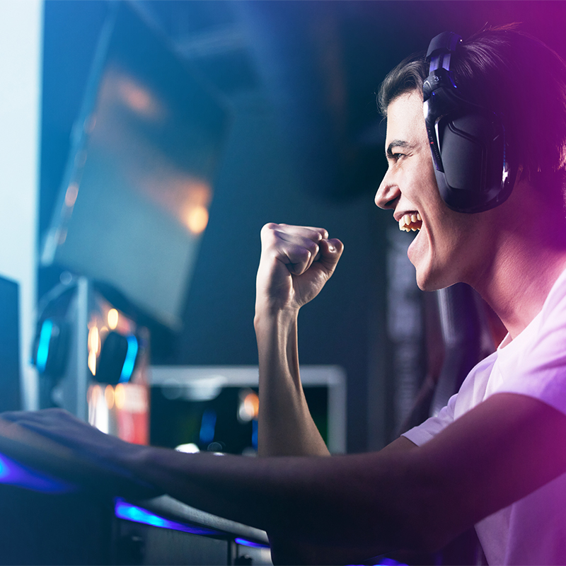 Giovane con cuffie esulta davanti a uno schermo. Il pugno � alzato in segno di vittoria. Luci blu e rosa illuminano la scena.