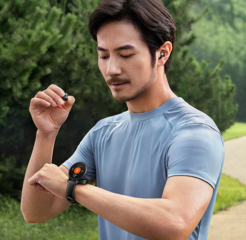 Uomo con maglietta blu guarda orologio da polso. Indossa auricolare; ne tiene un altro in mano. Sfondo verde con sentiero.