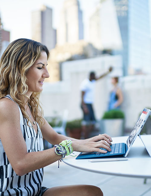 Donna che usa un laptop blu su un tavolo bianco, con persone e edifici sullo sfondo.