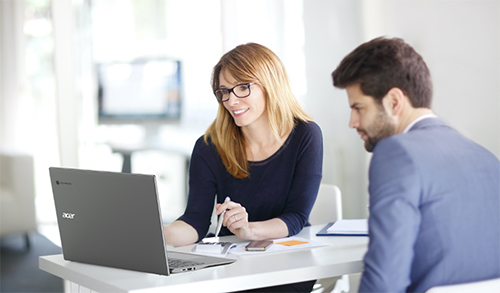 Donna e uomo seduti a una scrivania bianca con un laptop Acer, documenti e telefono. La donna sorride e indica lo schermo.