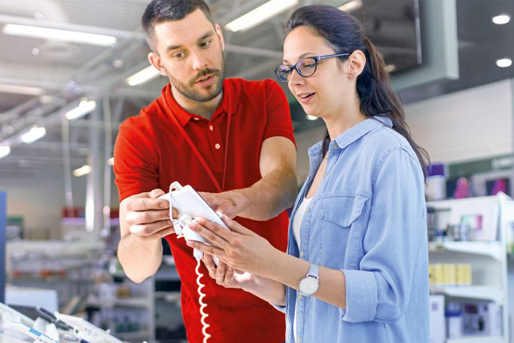 In un negozio, un uomo in rosso mostra un telefono bianco a una donna in azzurro. Scaffali sullo sfondo.