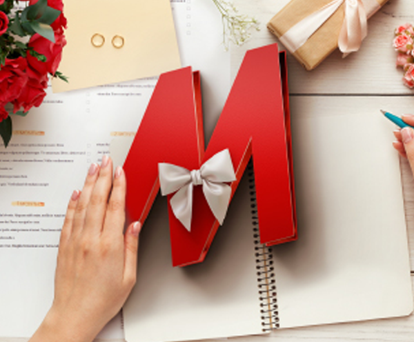 Lettera rossa "M" con fiocco su quaderno a spirale. Mani con penna e fiori completano la scena su sfondo di legno chiaro.
