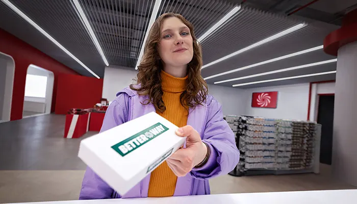 Donna con scatola bianca "Better Way" in ufficio moderno con accenti rossi e grigi. Scatole impilate sullo sfondo.