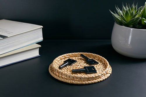 Tre chiavi nere su un sottobicchiere di vimini, accanto a libri impilati e una pianta in vaso grigio su una superficie scura.