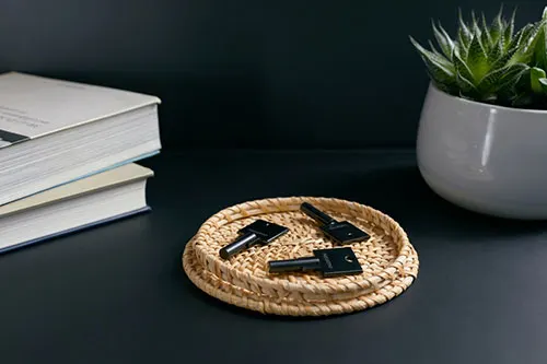 Tre chiavi nere su un sottobicchiere di vimini, accanto a libri impilati e una pianta in vaso grigio su una superficie scura.