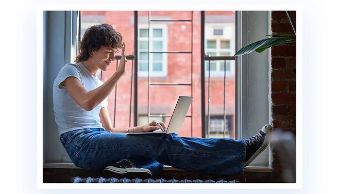 un ragazzo saluta verso il notebook mentre è seduto nei pressi di una finestra