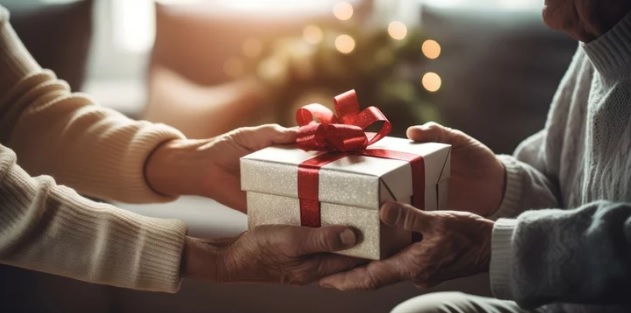 Due mani offrono una scatola regalo bianca con nastro rosso. Sfocati, divano e luci sullo sfondo.