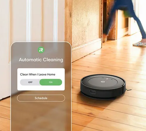 Aspirapolvere robot nero su pavimento in legno. Uno smartphone mostra l'impostazione "Pulizia automatica" con opzione "On".