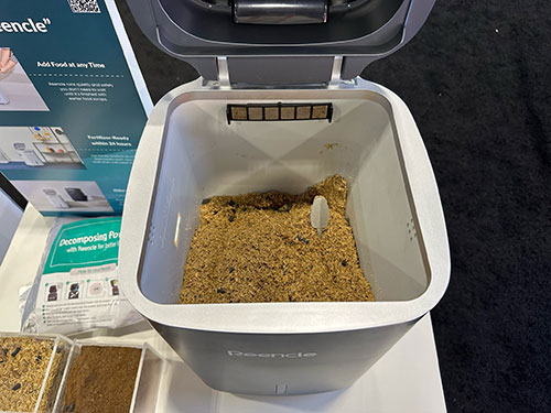 Contenitore bianco con coperchio aperto riempito di materiale organico marrone. Un cucchiaio bianco � visibile dentro.