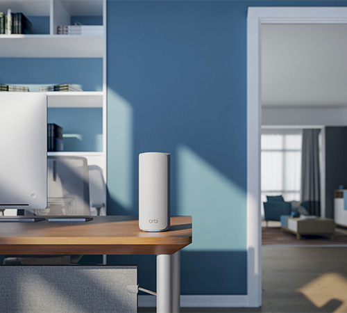 Scrivania di legno con cilindro bianco, monitor e scaffali. Muro blu e porta aperta su un soggiorno.