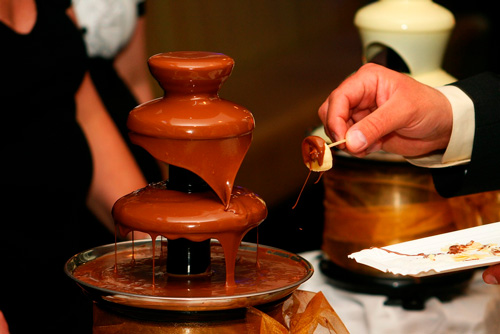 Fontana di cioccolato a pi� livelli con una mano che immerge una banana. Sullo sfondo, altre persone e attrezzature.