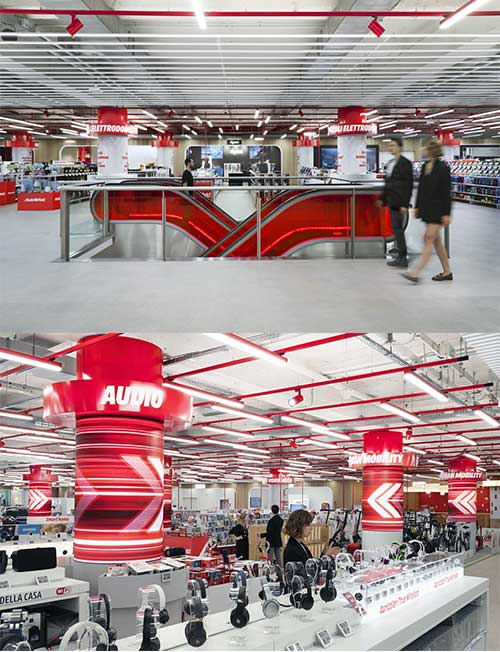 Immagine di un negozio con scaffali di prodotti, scale mobili rosse e pilastri con scritte rosse. Persone in movimento.