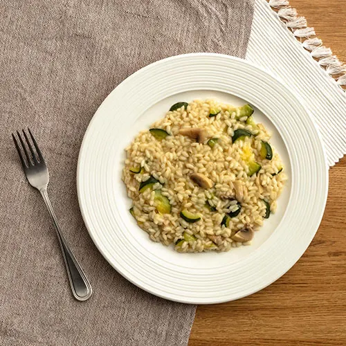 Piatto bianco con risotto alle zucchine e funghi, posizionato su una tavola di legno con tovaglioli e forchetta.