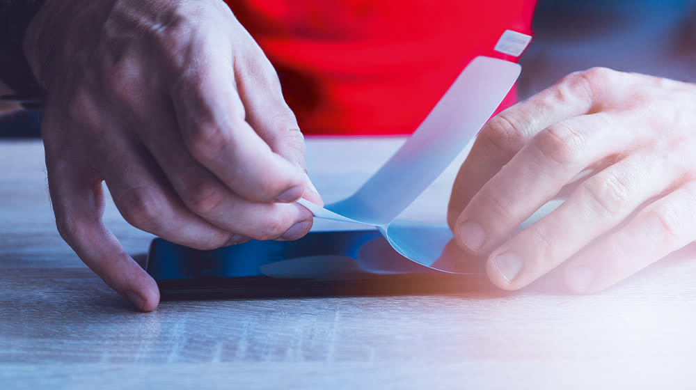 Due mani staccano una pellicola protettiva da uno smartphone blu su un tavolo chiaro, con sfondo rosso.