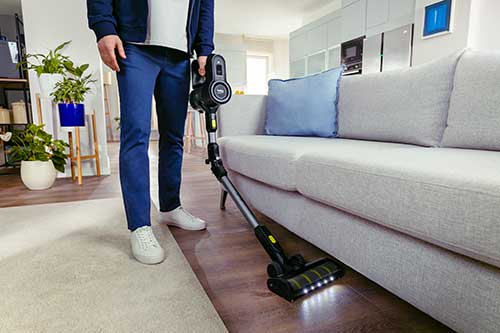 Un uomo aspira la polvere con un aspirapolvere nero in un soggiorno. C'� un divano chiaro, piante e scaffali in background.
