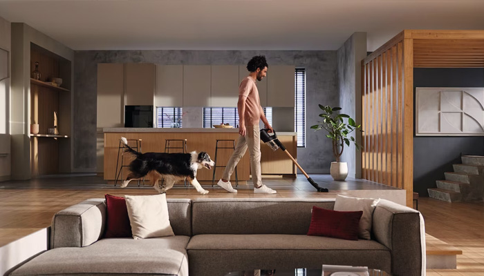 Uomo che usa l'aspirapolvere in salotto, con cane. Divano grigio in primo piano, cucina aperta sullo sfondo.