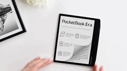E-reader "PocketBook Era" su tavolo bianco, con foto incorniciata e fiori sullo sfondo. Mani che sfogliano lo schermo.