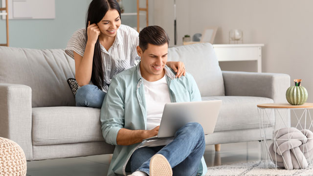 Uomo seduto sul pavimento usa un laptop, donna seduta sul divano dietro di lui. Soggiorno con divano grigio chiaro e tavolino.