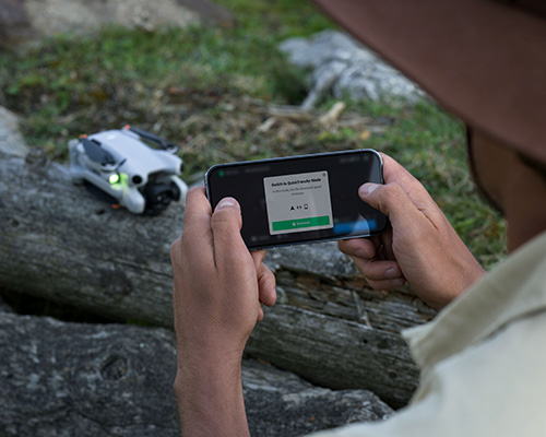 Uomo che tiene un telefono con un'app di volo; sullo sfondo, un piccolo drone bianco su una roccia.