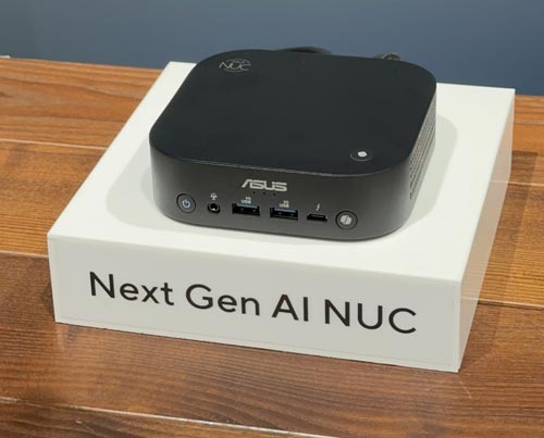 Un piccolo computer nero ASUS è posto su una scatola bianca con la scritta "Next Gen AI NUC" su un tavolo di legno.