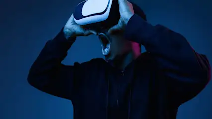 Un uomo con felpa nera usa visore VR bianco. Ha la bocca aperta, sfondo blu scuro con luci rosa.