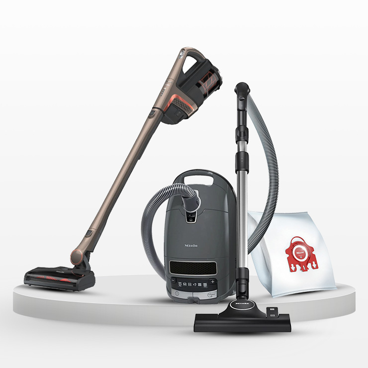 Tre aspirapolveri di colore diverso su una piattaforma bianca con un sacchetto di carta.