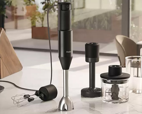 Un frullatore a immersione nero con accessori su un ripiano bianco. Include frusta, tritatutto e base. Sullo sfondo, piante e sedie.