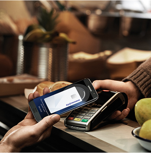 Mani che tengono un telefono con Samsung Pay vicino a un terminale di pagamento, con cibo e contenitori sullo sfondo.