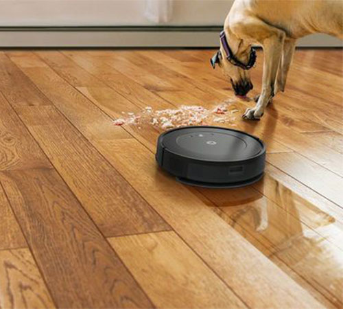 Su un pavimento in legno, un robot aspirapolvere nero � vicino a dei croccantini per cani sparsi. Un cane si china per mangiarli.