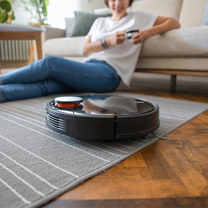 Un aspirapolvere robot nero su un tappeto grigio a righe bianche; una persona seduta su un divano sullo sfondo.