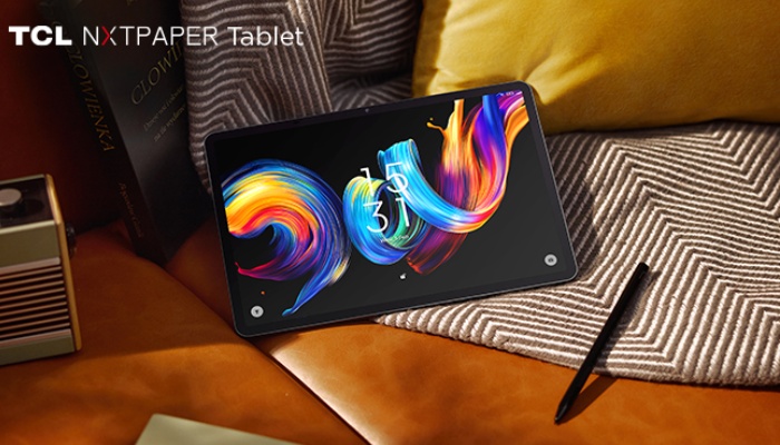 Un tablet TCL NXTPAPER Tablet appoggiato su un divano in pelle accanto a cuscini e a una penna stilo. Lo schermo mostra uno sfondo nero con onde fluide e colorate e l'orario digitale 15:31.