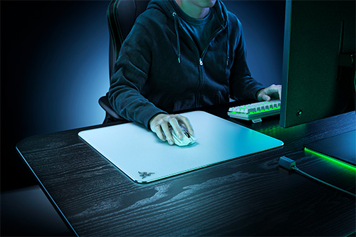 Uomo con felpa con cappuccio seduto alla scrivania. Utilizza mouse su tappetino bianco, tastiera e monitor visibili. Luce blu e verde.